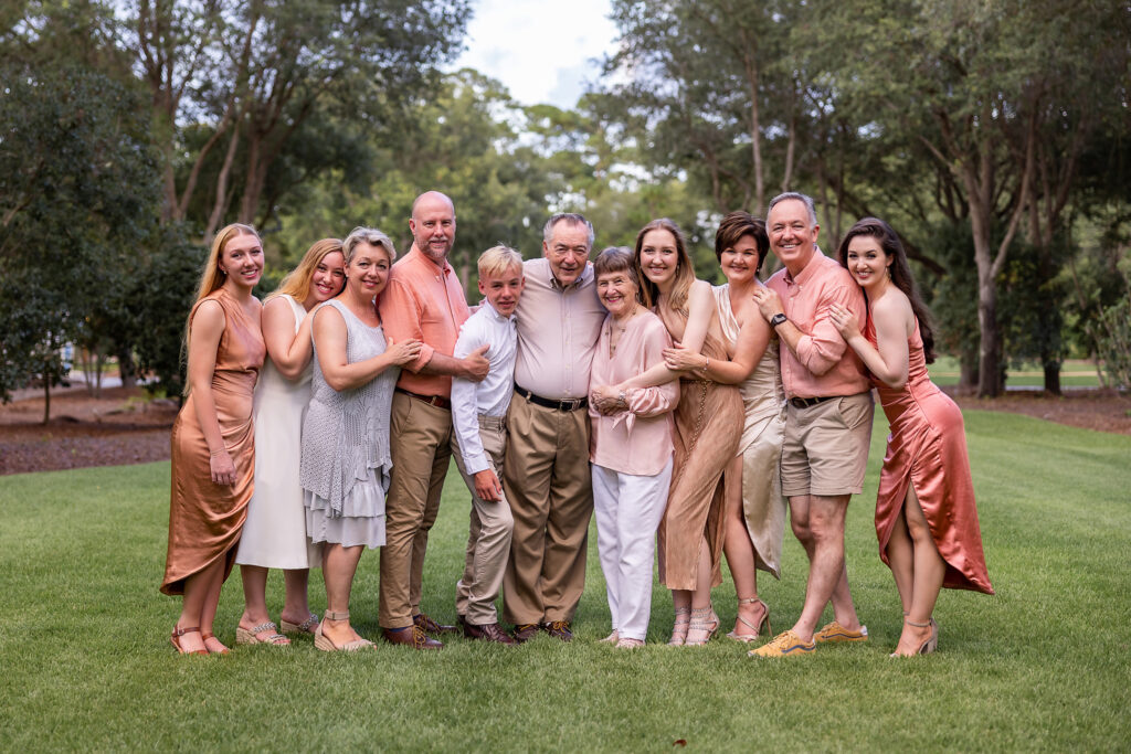 A family of 11 posing for a portrait at Watercolor wearing shades of peach and earthy coral mixed with khaki.