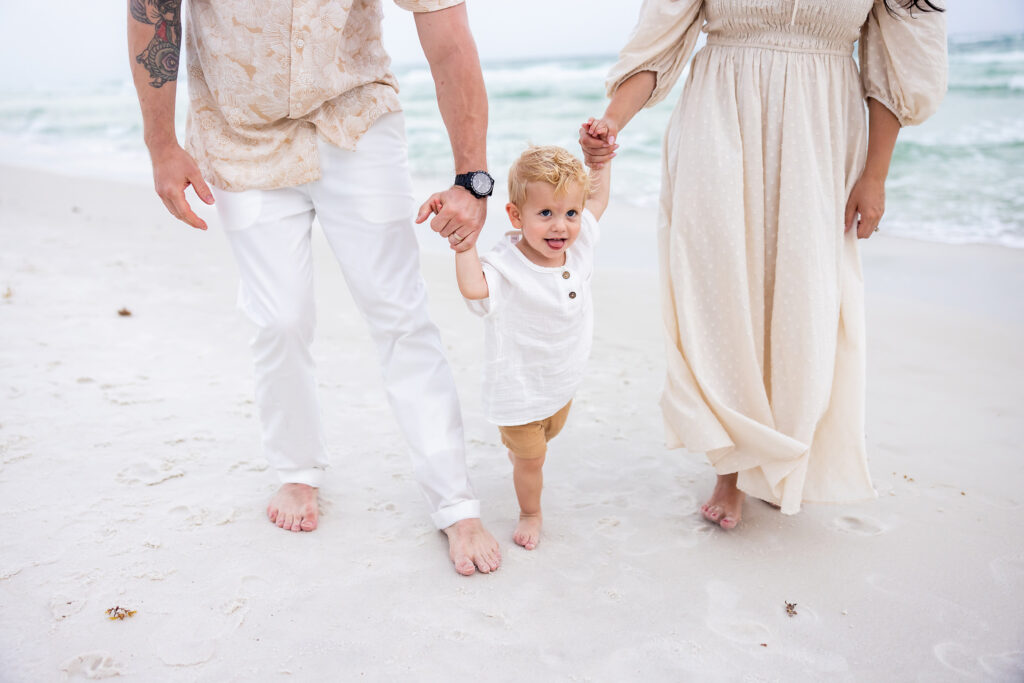 Mom and dad holding the hand of a toddler boy walking on the beach at Seacrest on 30a and they are wearing white and creams.
