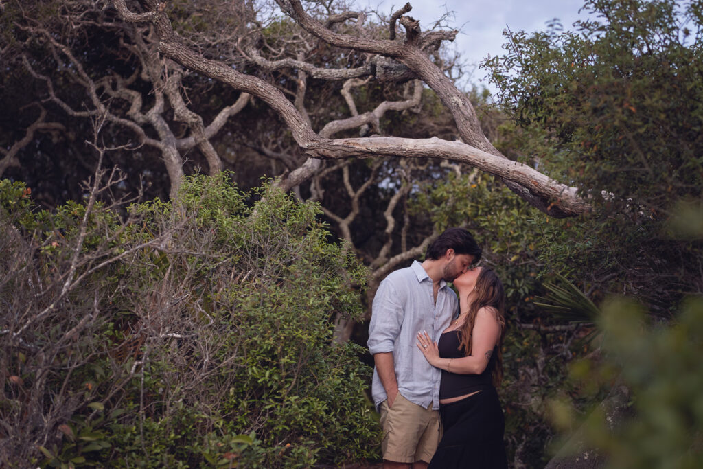 kissing photo of newly engaged couple in front of the heavy tree area of the trail at Grayton Beach State Park.