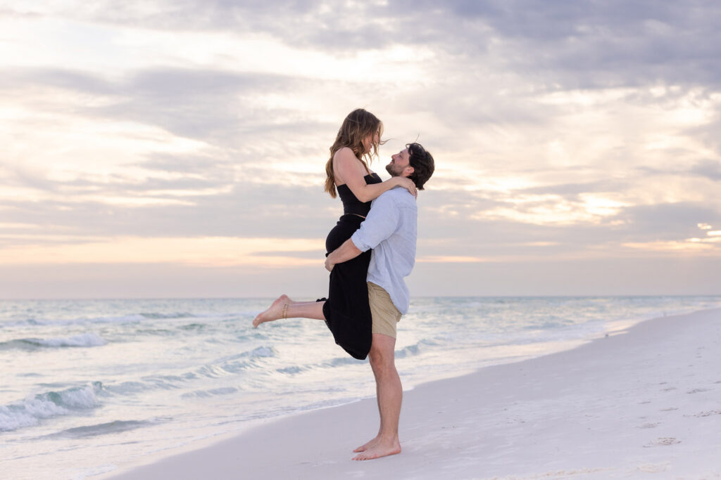 man holding woman in air on beach after proposing.