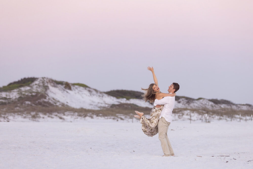 And excited shot of the new fiance kicking her feet back while being picked up by her boyfriend with dunes in the background at sunset on 30a.